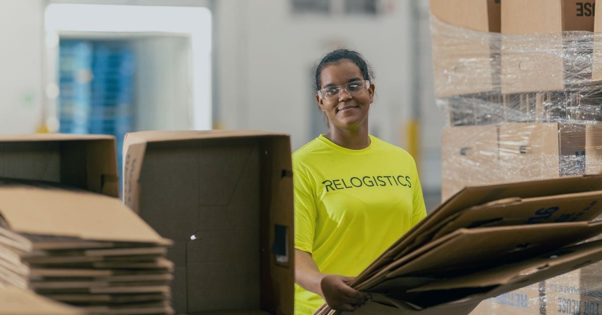 Woman in a bright yellow Relogistics shirt handling cardboard boxes in a distribution center, emphasizing salvage operations and efficient logistics processes.