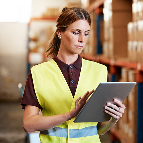 Woman in yellow safety vest using tablet in a warehouse, emphasizing supply chain efficiency for e-commerce logistics.