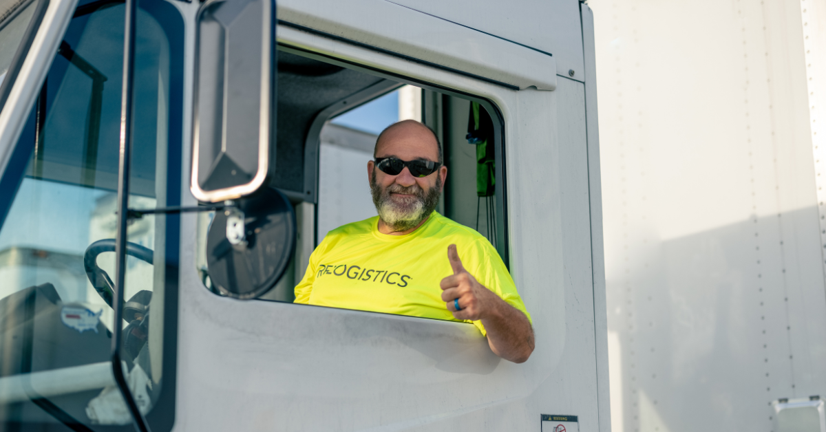 Smiling truck driver in a bright yellow Relogistics shirt giving a thumbs-up from the cab of a delivery truck, representing efficient supply chain management and customer service.