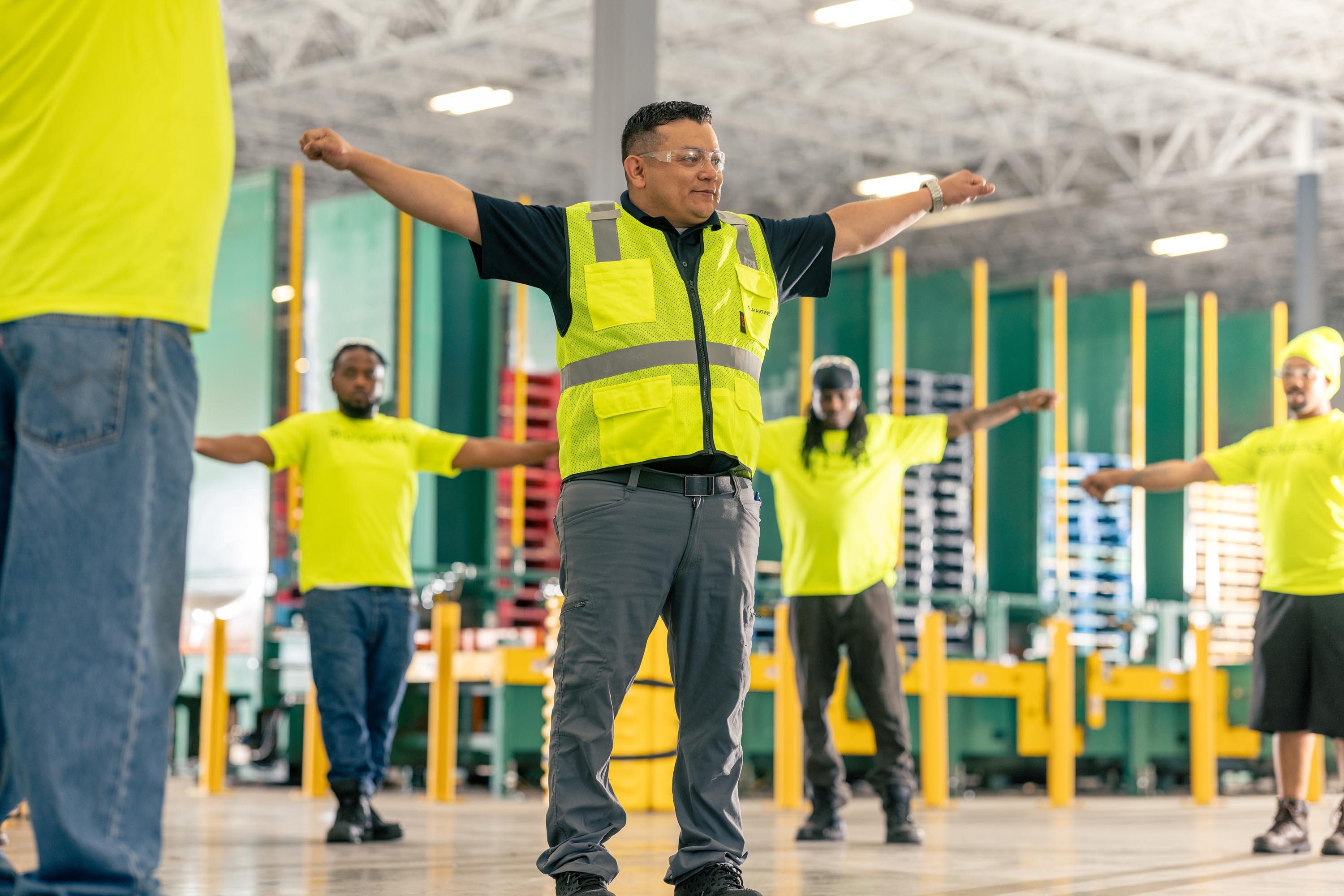 Warehouse employees participating in a safety training session, wearing bright yellow safety vests, demonstrating stretching exercises to promote workplace safety and health.
