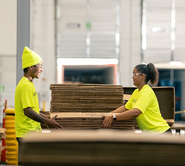 Two warehouse workers in bright yellow shirts and safety gear collaborating to handle and stack cardboard boxes, emphasizing labor efficiency in supply chain management.
