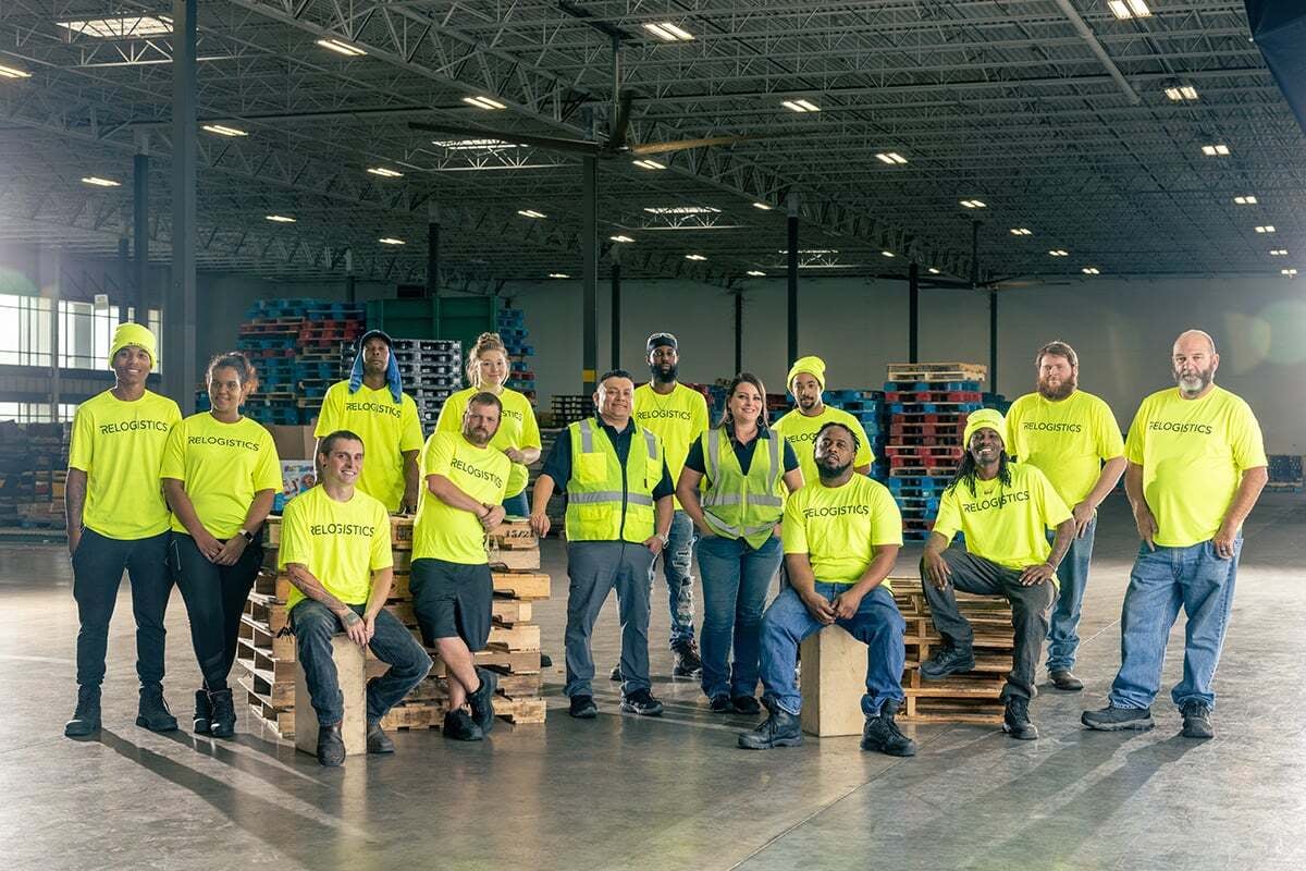 Group of Relogistics employees in bright yellow shirts and safety vests posing in a warehouse, showcasing teamwork in pallet management and supply chain efficiency.