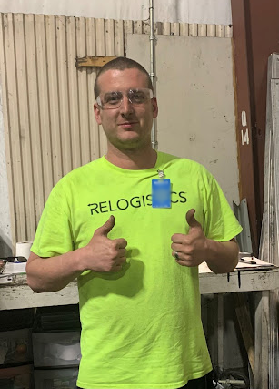 Man in a bright yellow safety shirt with "RELOGISTICS" logo, giving thumbs up in a warehouse setting, promoting safety and positivity in logistics operations.