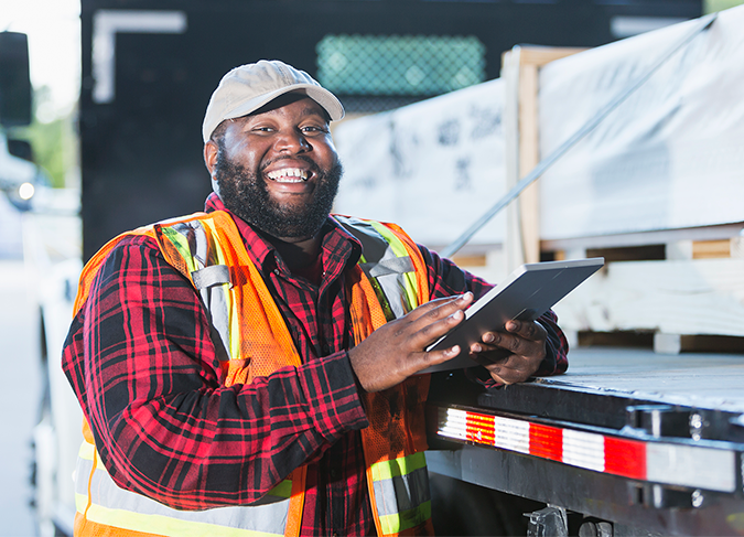 Smiling logistics worker in safety vest using tablet on truck, representing supply chain efficiency and operational support for e-commerce solutions.