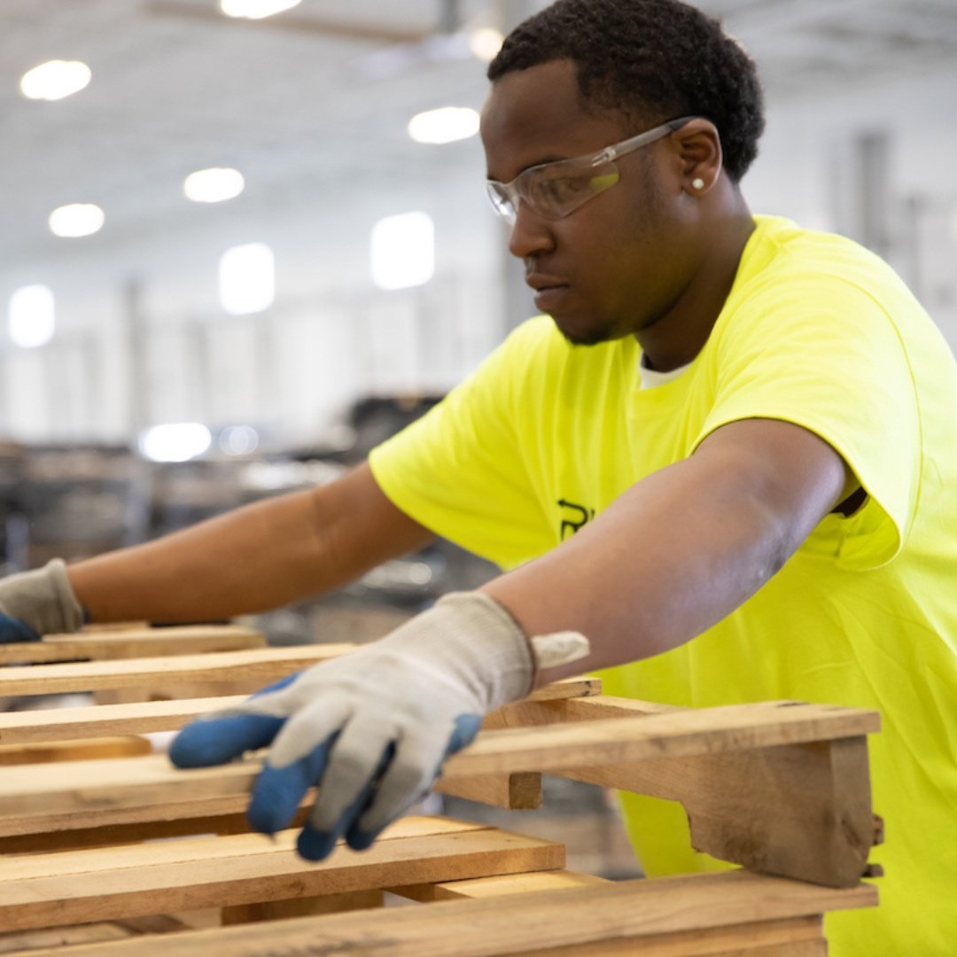 Worker in bright yellow safety shirt and gloves assembling wooden pallets in a warehouse, emphasizing commitment to safety in operations.
