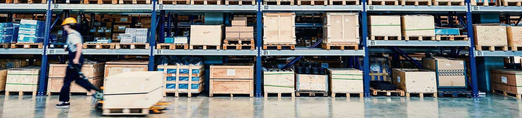 Warehouse worker moving a pallet jack among stacked boxes and pallets in a logistics facility, emphasizing efficient warehouse operations and support activities.